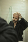 Man with thinning hair and a balding crown examining his scalp in the bathroom mirror, the daily concern that drives men to seek natural hair growth solutions like Medité