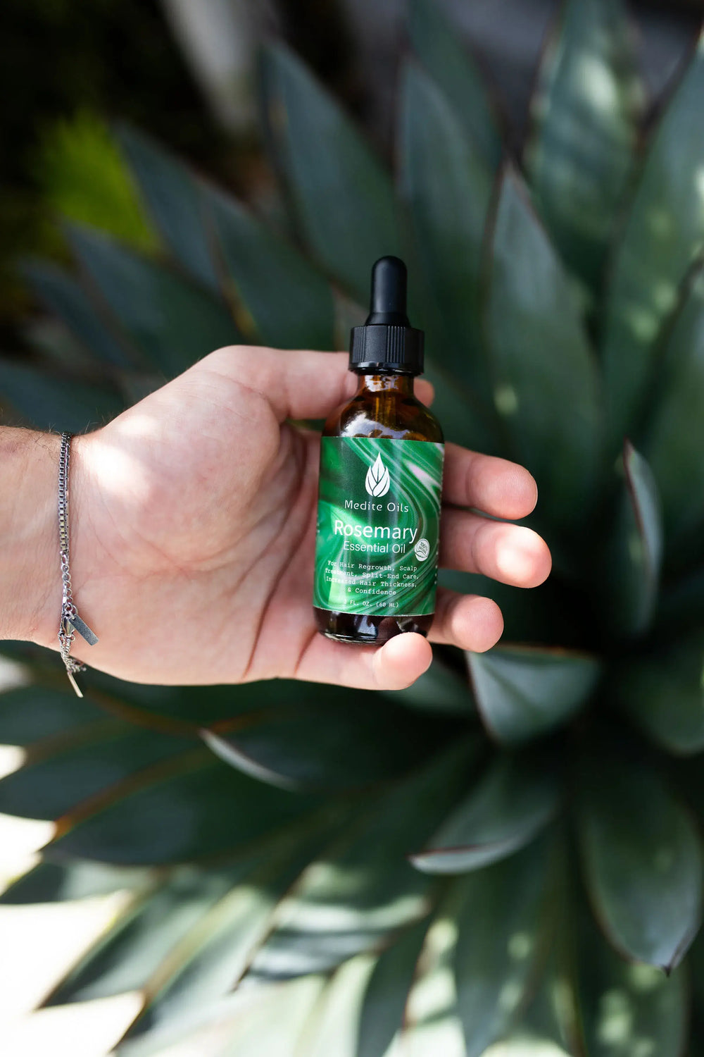 Hand holding amber bottle of Medité plant-based Rosemary Essential Oil for hair growth and scalp care in front of fresh green plants