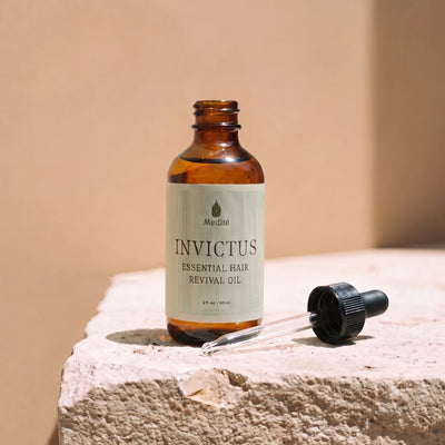 An amber glass dropper bottle labeled 'Medité Invictus Essential Hair Revival Oil' sits on a textured stone surface with its black dropper cap placed beside it against a warm beige background.